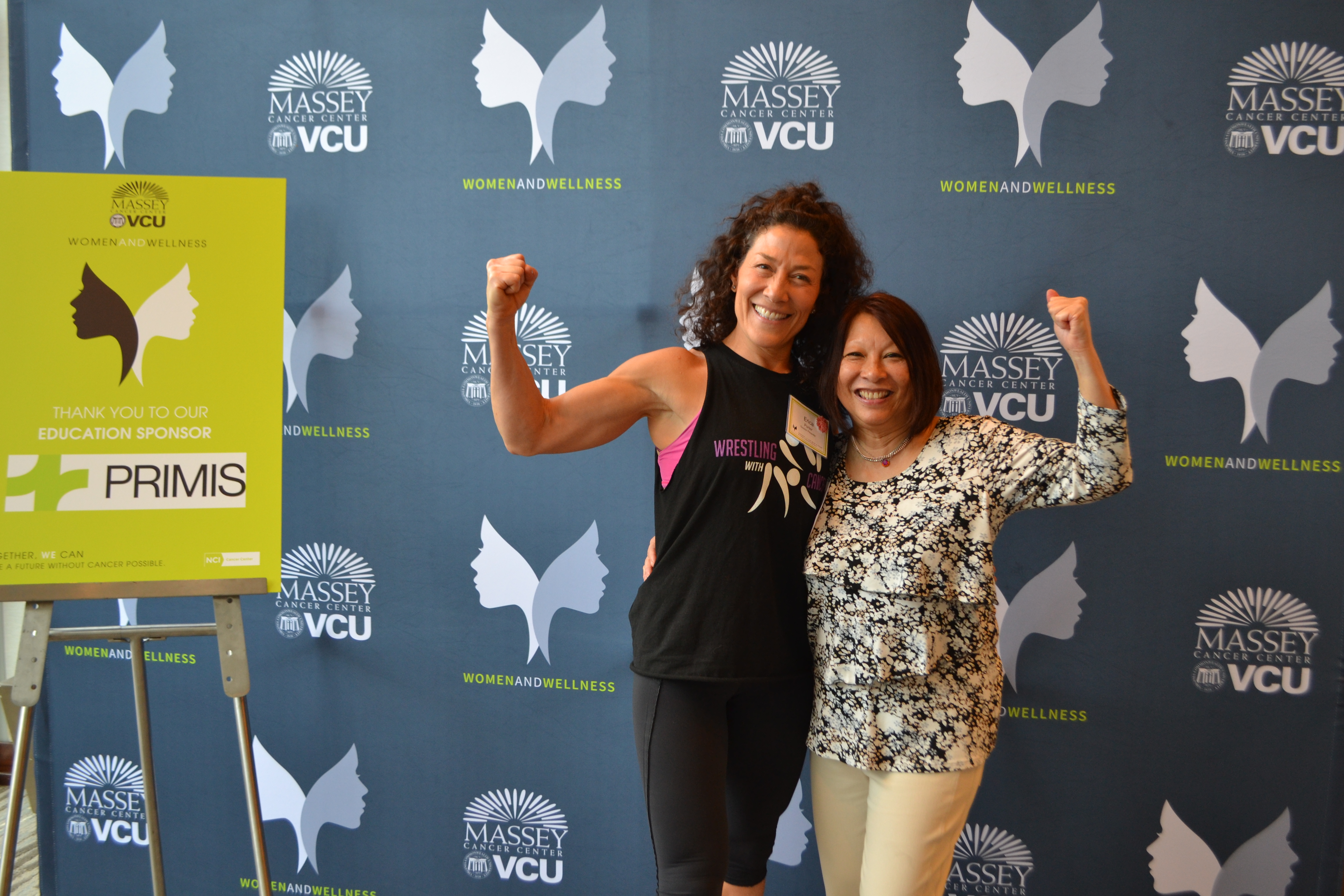 Two woman hugging and using their opposing arm to do a flex motion in front of Women and Wellness-branded backdrop